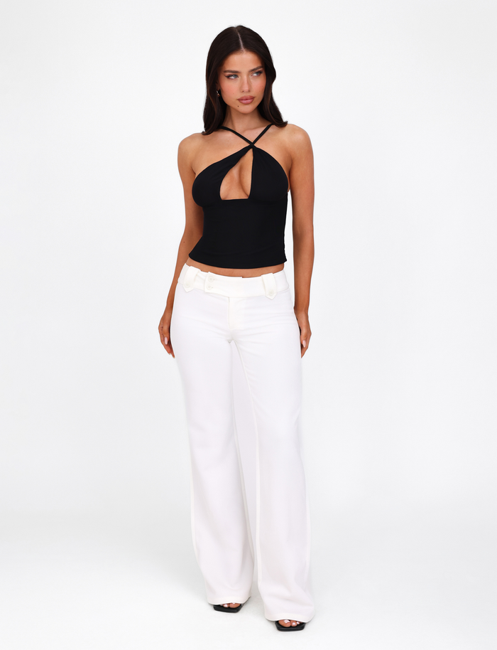 A woman with long dark hair models the TIGERMIST ACADIA TOP in black, featuring an asymmetric neckline, paired with high-waisted white wide-leg pants. She stands against a plain white background, gazing slightly to the side with her hands at her sides.