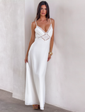A woman with long dark hair models the AISLYNN MAXI DRESS - WHITE by TIGERMIST, a white slip maxi dress with lace details, posing gracefully against a light-colored wall with one hand on her hip.