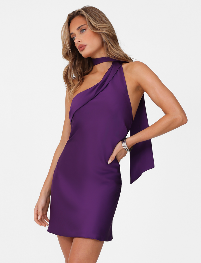 A woman poses against a white background wearing the TIGERMIST ALLIRA MINI DRESS 2.0 - PURPLE : LAVENDER, a sleeveless asymmetrical satin dress with a matching neck scarf, looking to the side with one hand on her hip.