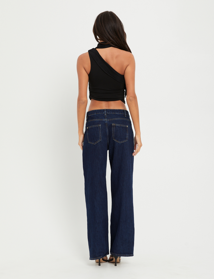 A woman with long dark hair faces away, wearing the TIGERMIST ALLIRA TOP in black—an asymmetrical, stretchy one-shoulder style—paired with dark blue wide-leg jeans and heels, set against a plain white background.