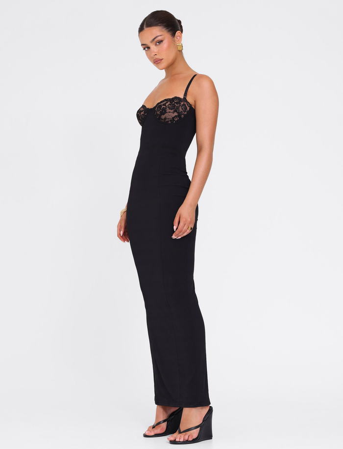 A woman stands against a white background wearing the TIGERMIST ALLURA MAXI DRESS in black, paired with black heels and gold earrings. Her hair is styled in a sleek bun, and she faces slightly to the side.