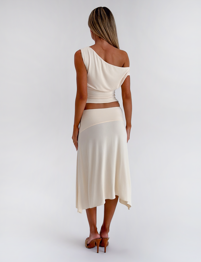A woman stands facing away, wearing the TIGERMIST ALYSSA HI LO SKIRT in cream with an asymmetrical hem and front leg slit, paired with a matching cream one-shoulder top and heeled sandals against a plain white background.