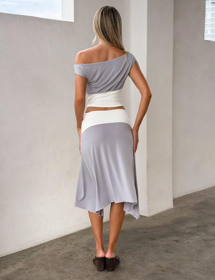 A woman indoors, facing away, wears the TIGERMIST ALYSSA HI LO SKIRT in grey with an asymmetrical hem and front slit, paired with a light gray off-shoulder top, white waist wrap, and brown sandals.
