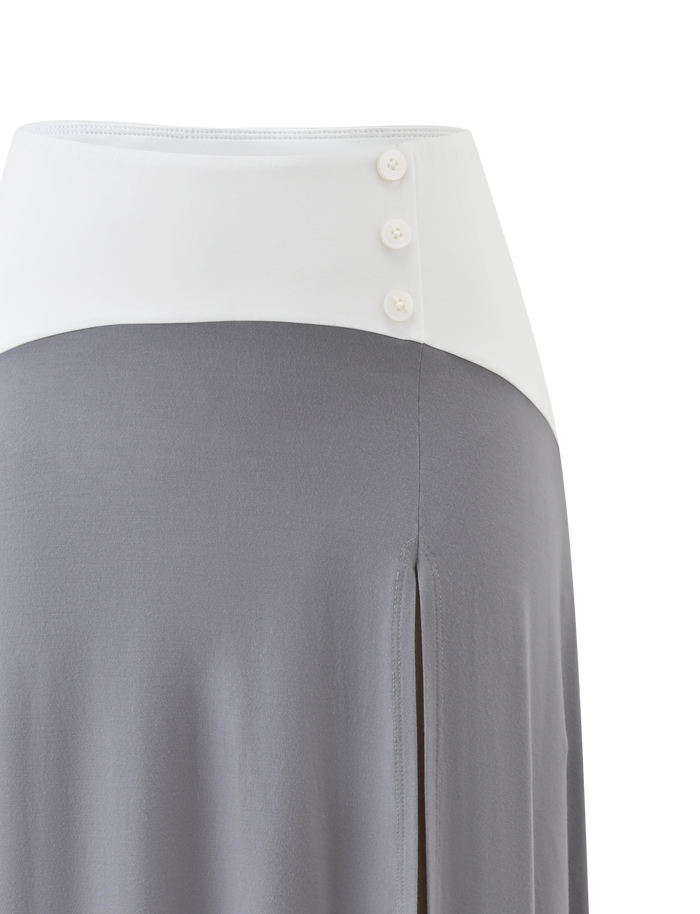 The ALYSSA HI LO SKIRT by TIGERMIST features a high waist, wide white waistband with three buttons, flowing grey fabric, asymmetrical hem, and a chic front leg slit.