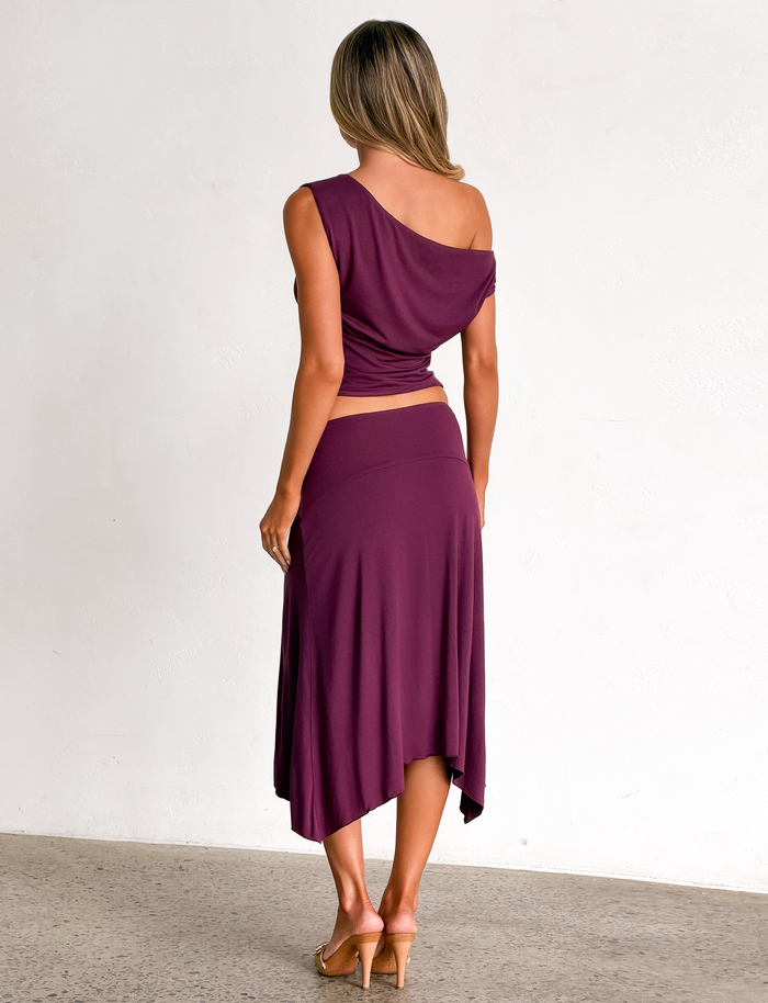 A woman is seen from behind wearing a purple one-shoulder top with the TIGERMIST ALYSSA HI LO SKIRT in plum, featuring an asymmetrical hem.