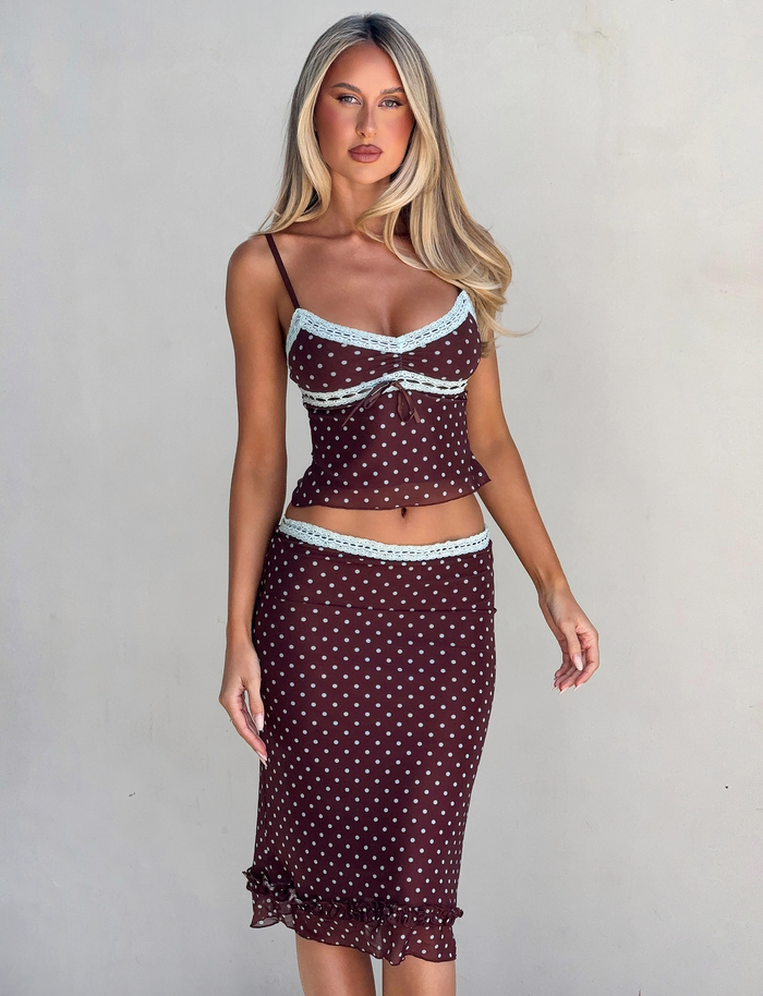 A woman with long blonde hair wears the TIGERMIST AMARA MIDI SKIRT - POLKA DOT set, featuring a fitted cami top and a ruffled-hem midi skirt with light blue lace trim, standing against a plain light background.