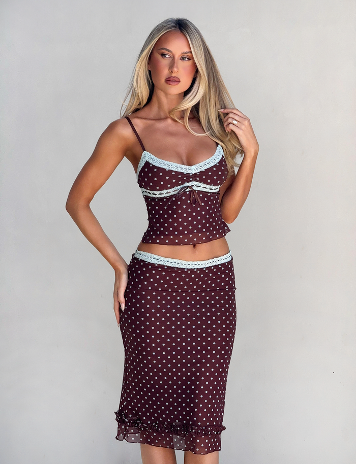 A woman with long blonde hair wears the TIGERMIST AMARA TOP - POLKA DOT, a brown sleeveless cami with light blue trim, paired with a matching skirt, standing against a plain light gray background.