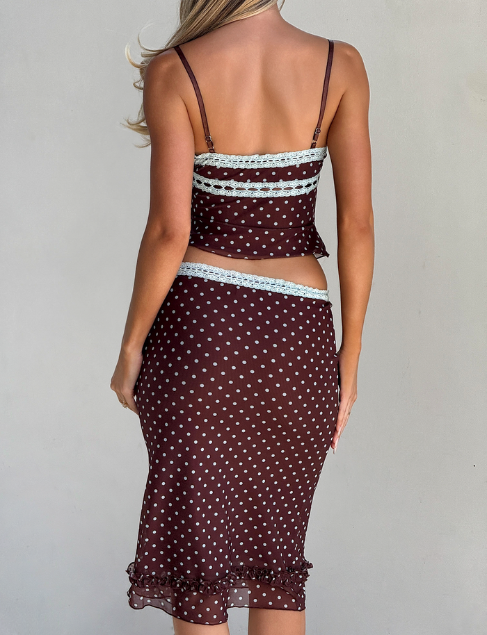 A woman with long blonde hair is seen from the back wearing the TIGERMIST AMARA TOP - POLKA DOT, a sheer brown cami with white dots, lace trim, and ruffle hem details for a feminine touch.