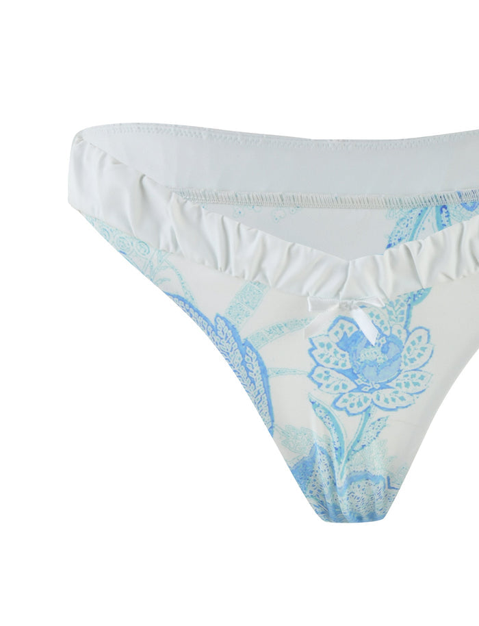 A detailed view of the AMARAN BIKINI BOTTOM - BLUE: FLORAL TAPESTRY by TIGERMIST highlights its white base adorned with a blue floral pattern. The fabric is elegantly accentuated with a subtle ruffle trim along the waistband, embodying a delicate and feminine aesthetic. The intricate floral motifs add an artistic touch that complements its elegant style.