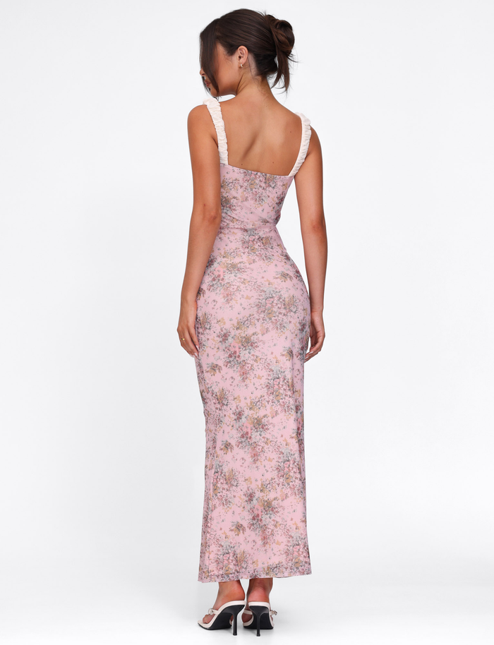 A woman, hair in a loose bun, wears the TIGERMIST AMARAN DRESS - PINK: CAMO FLORAL, a bodycon maxi with white ruffle straps, styled with black heeled sandals against a plain white background.