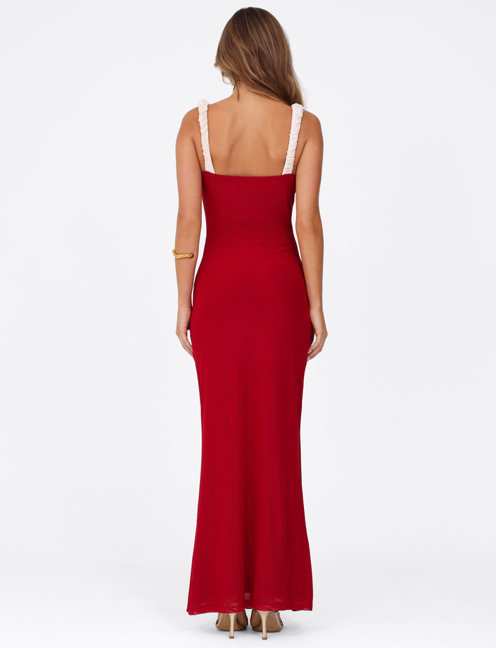 A woman with long hair, facing away, wears the TIGERMIST AMARAN MAXI DRESS - RED : BURGUNDY. The floor-length, sleeveless dress features light straps and is paired with beige heels against a plain white background.