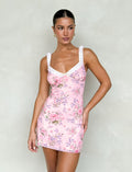 A woman wearing the AMARAN MINI DRESS - PINK : FLORAL : WILD FLOWER by TIGERMIST poses against a plain, light-colored background. Her hair is styled in a sleek bun, and she accessorizes with hoop earrings.