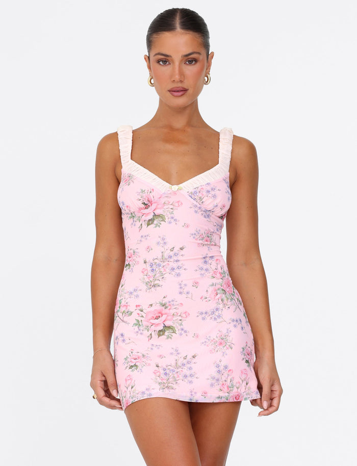 A woman wearing the AMARAN MINI DRESS by TIGERMIST in a pink wild flower print with bustier styling and ruffled straps poses against a plain white background. Her hair is tied back, and she accessorizes with hoop earrings while she gazes directly at the camera with a neutral expression.