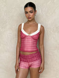 A person stands against a textured beige wall wearing a TIGERMIST AMARAN TOP - RED CHECK, featuring a white ruffled neck detail. They have slicked-back hair, hoop earrings, and a calm expression.