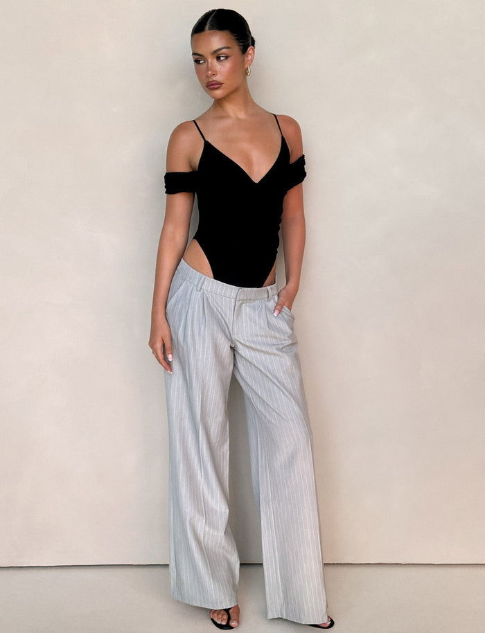 A woman stands against a light beige wall, wearing TIGERMIST's AMBERLY SUIT PANT in grey pinstripe and a black cold-shoulder bodysuit. Her hair is tied back, one hand in her pocket as she looks to the side.