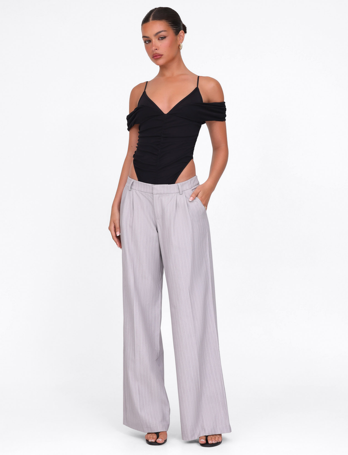 A woman wearing TIGERMIST’s AMBERLY SUIT PANT in grey pinstripe stands against a white background, hands in her pockets. She pairs the tailored wide-leg pants with a black off-shoulder top, open-toe heels, and has her hair tied back.