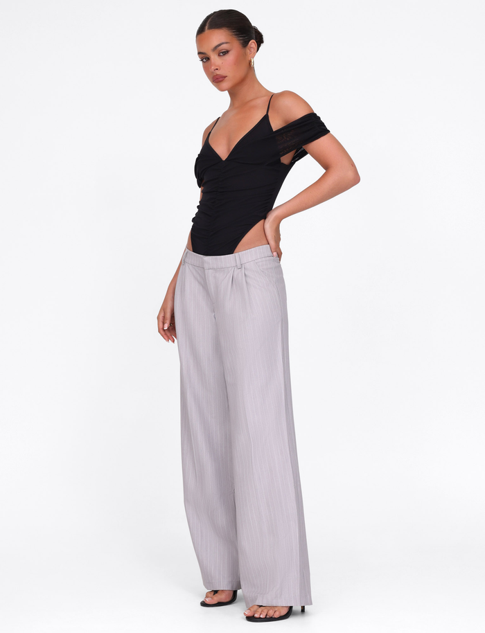 A woman poses against a white background, wearing TIGERMIST’s AMBERLY SUIT PANT in grey pinstripe with a black off-shoulder top and black open-toe heels. She stands with one hand on her hip, hair styled in a bun.