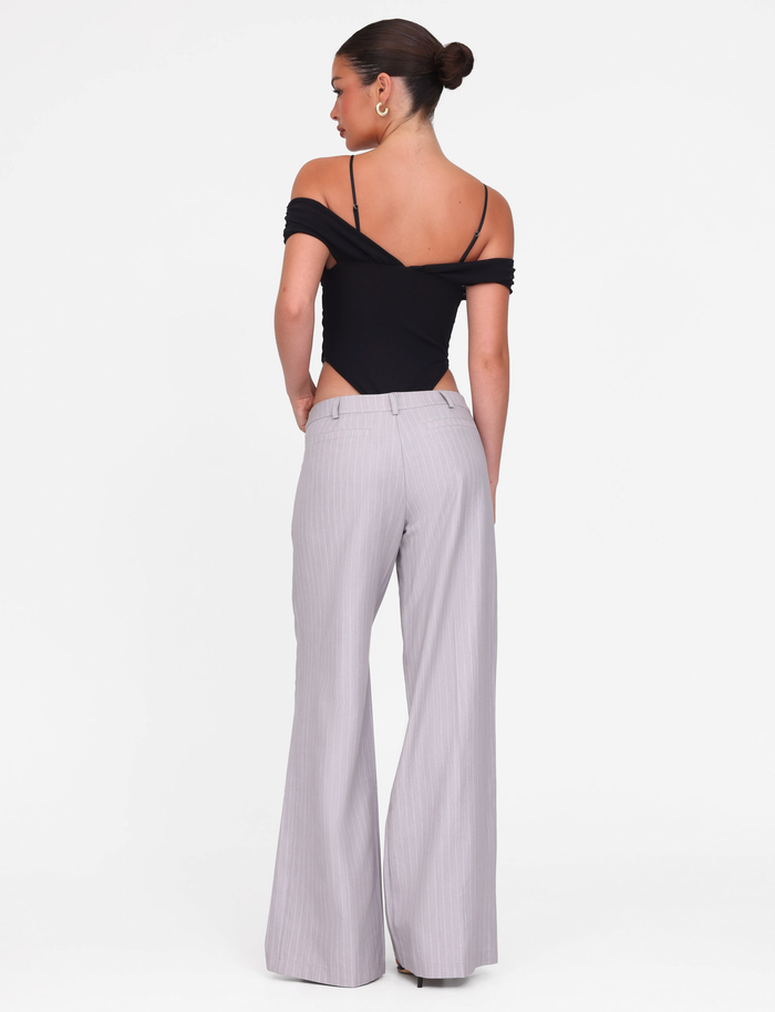 A woman with her hair in a bun stands against a plain white background, wearing TIGERMIST’s AMBERLY SUIT PANT in grey pinstripe and a black off-shoulder top.