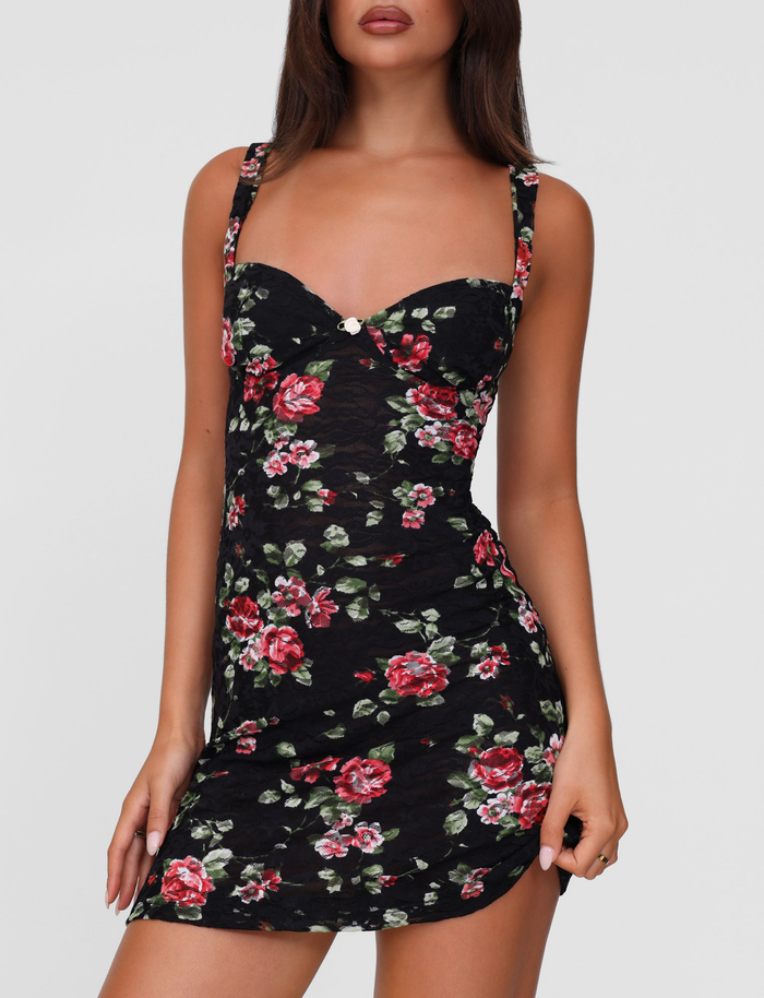 A woman wears the TIGERMIST ANDEE MINI DRESS in BLACKROSE, a fitted, sleeveless stretch lace dress with thin straps, a sweetheart neckline, and red and pink rose florals on a light background.