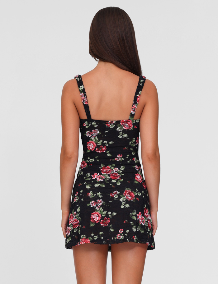 A woman with long brown hair stands with her back to the camera, wearing the TIGERMIST ANDEE MINI DRESS in Blackrose—a sleeveless dress featuring red and green floral prints—against a plain, light-colored background.