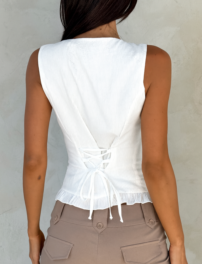 A woman is seen from behind wearing the TIGERMIST ANIYA KEYHOLE TOP in white, featuring a sleeveless design and lace-up back, paired with high-waisted beige pants with pockets against a plain light-colored wall.