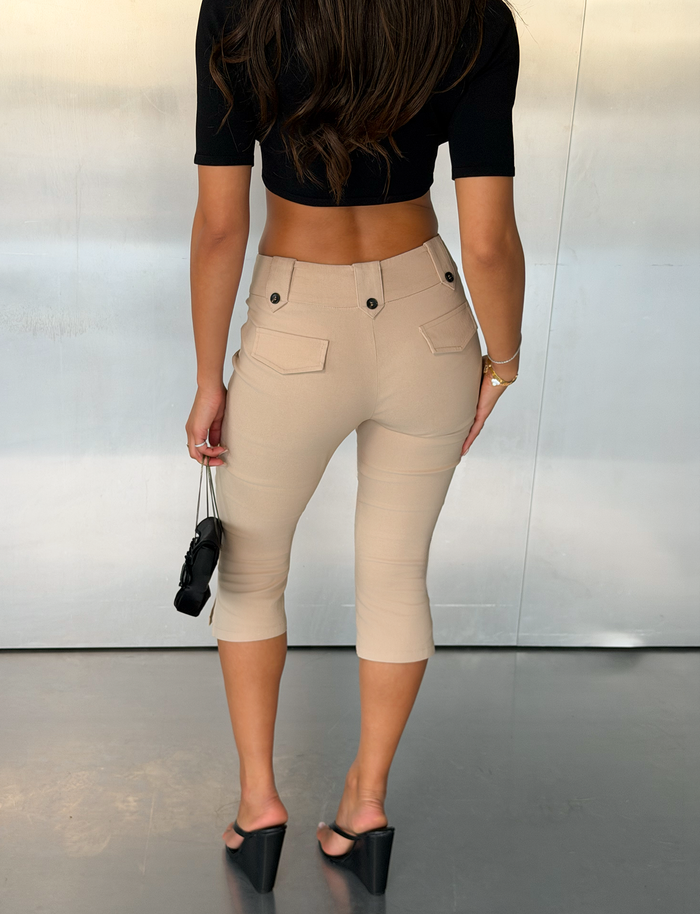 A woman stands indoors facing away, wearing a black crop top, the TIGERMIST ARIA CAPRI - KHAKI tailored fit mid-rise capri pants, and black wedge sandals, holding a small black purse in her left hand.
