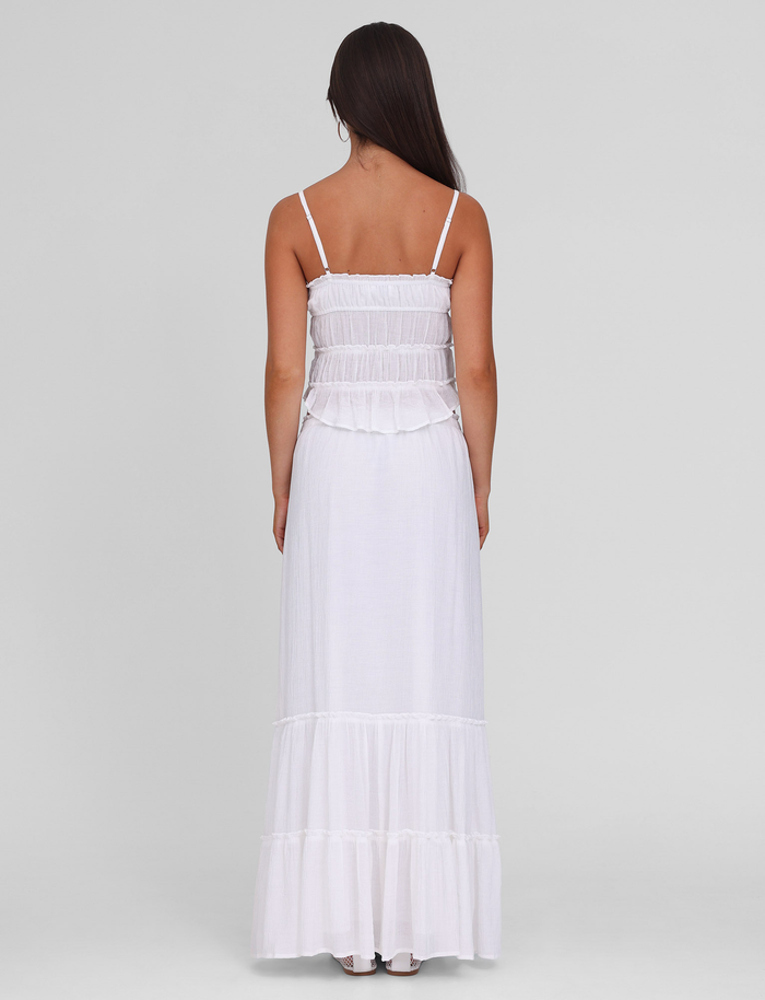 A woman with long brown hair, seen from behind, wears the TIGERMIST ASHLYNN MAXI SKIRT in white, featuring an elasticated waist and ruffle detailing. She stands against a plain light gray background.