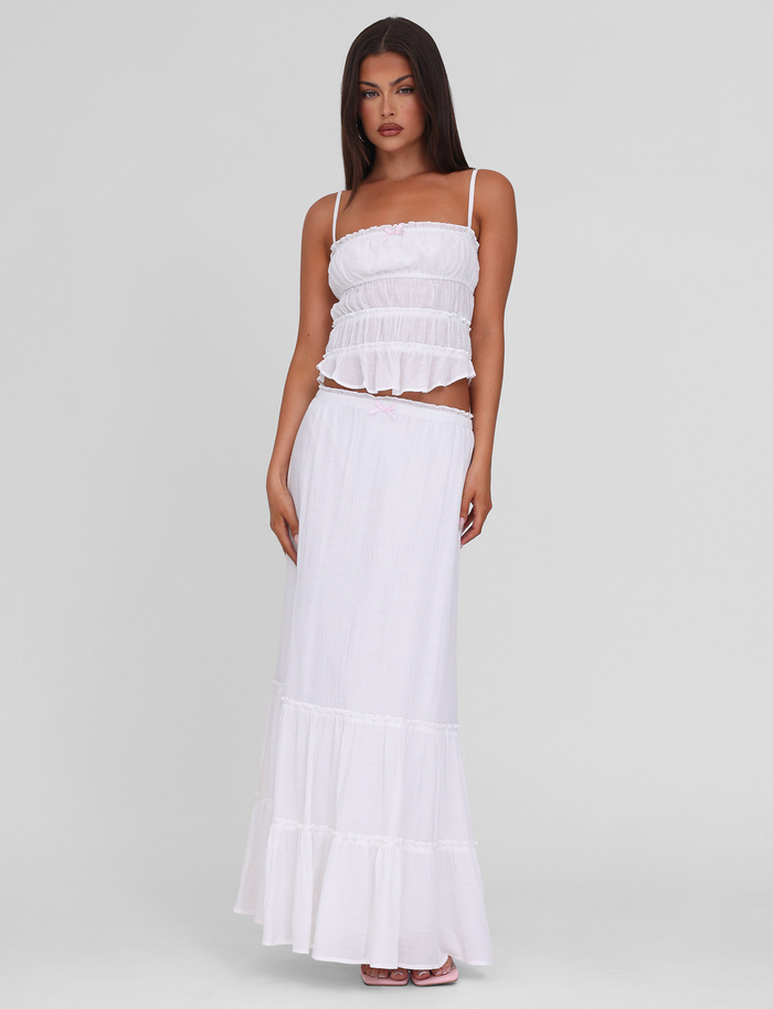 A woman with straight dark hair and a neutral expression wears the TIGERMIST ASHLYNN TOP in white—a sleeveless, tiered cami crop top with an elastic underbust—paired with a long, flowing white skirt against a plain light background.