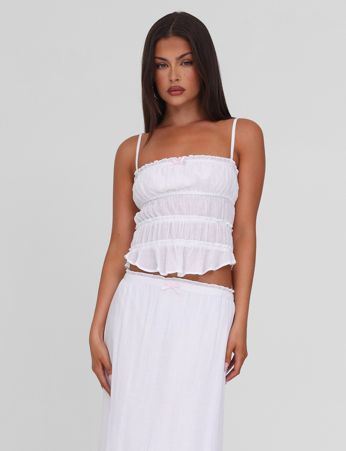 A woman with long dark hair models the TIGERMIST ASHLYNN TOP - WHITE, a tiered cami with adjustable straps, paired with a matching long white skirt against a plain light gray background.