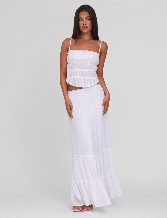 A woman stands against a plain background wearing the TIGERMIST ASHLYNN TOP in white, featuring adjustable shoulder straps. She pairs it with a matching flowing skirt, has long dark hair, and looks confidently at the camera with neutral makeup.