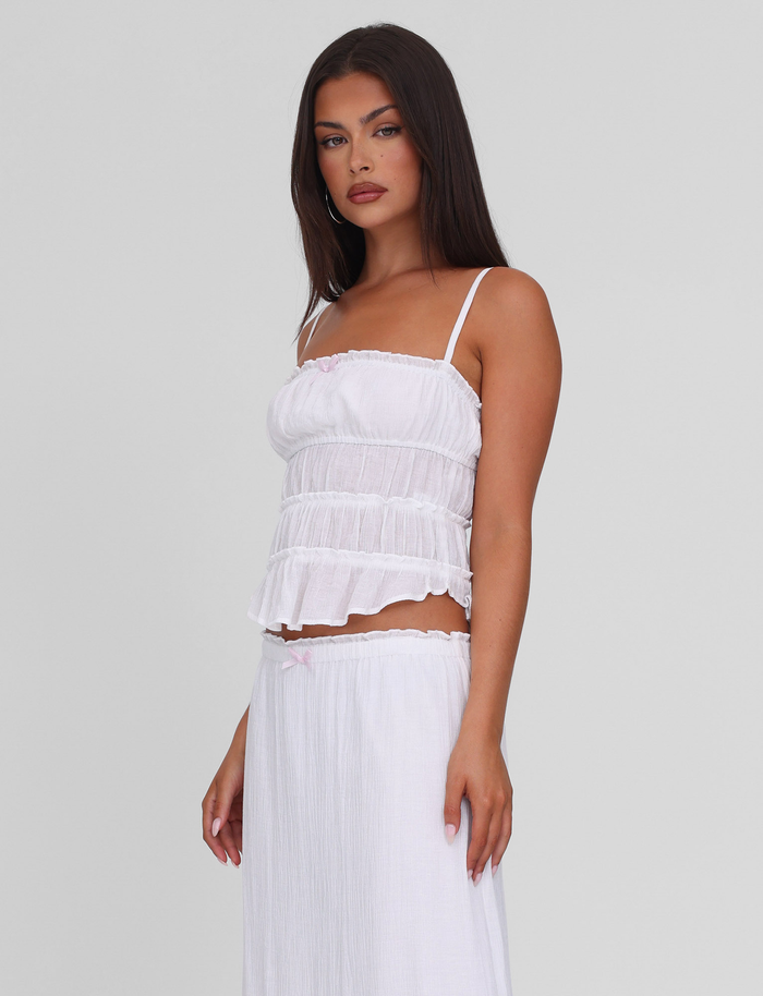 A woman with long dark hair wears the TIGERMIST ASHLYNN TOP in white, featuring adjustable straps. She pairs it with a matching skirt and stands against a plain light gray background, gazing slightly to the side with a neutral expression.