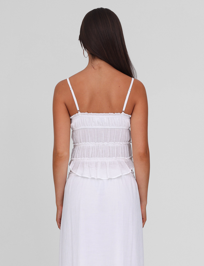 A woman with long brown hair faces away, wearing the TIGERMIST ASHLYNN TOP in white, featuring adjustable straps and a ruffled, textured bodice, set against a plain light gray background.