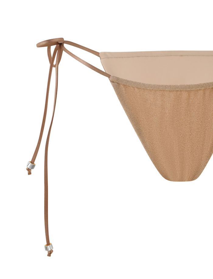 Close-up of the TIGERMIST AVE BIKINI BOTTOM - GOLD, showcasing adjustable side ties with metallic accents. The beige fabric features a subtle gold sparkle for a chic, shimmery touch.