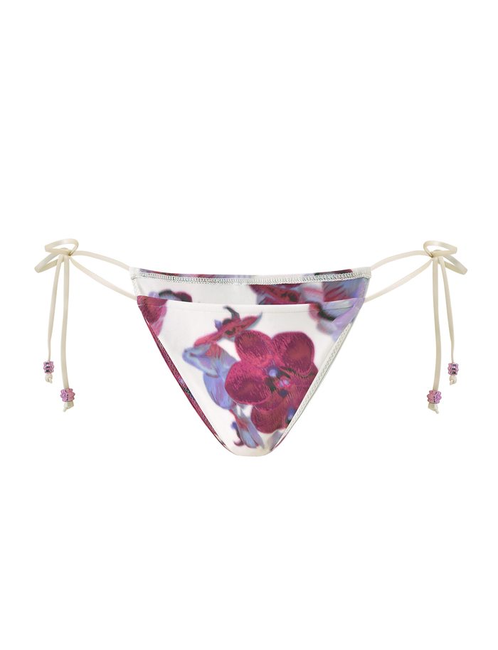The AVE BIKINI BOTTOM - ORCHID HAZE by TIGERMIST is a cheeky, side-tie bikini bottom with a white base and bold pink and purple floral print. String ends are accented with playful beads.