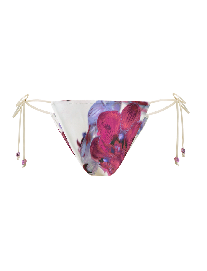 The TIGERMIST AVE BIKINI BOTTOM - ORCHID HAZE features a cheeky cut with white side ties adorned with floral beads, and displays a vibrant pink and purple flower print on a crisp white background.