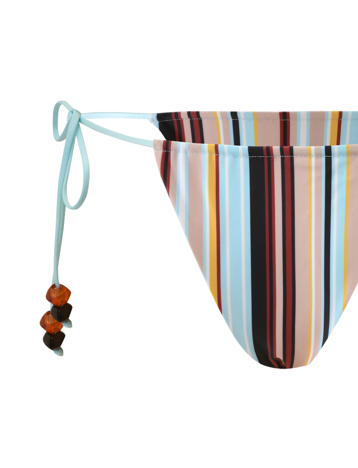 A close-up of the TIGERMIST AVE BIKINI BOTTOM - VARIEGATED STRIPE highlights its cheeky fit, adjustable side ties with brown beads, and bold vertical stripes in blue, black, red, orange, and beige on white.