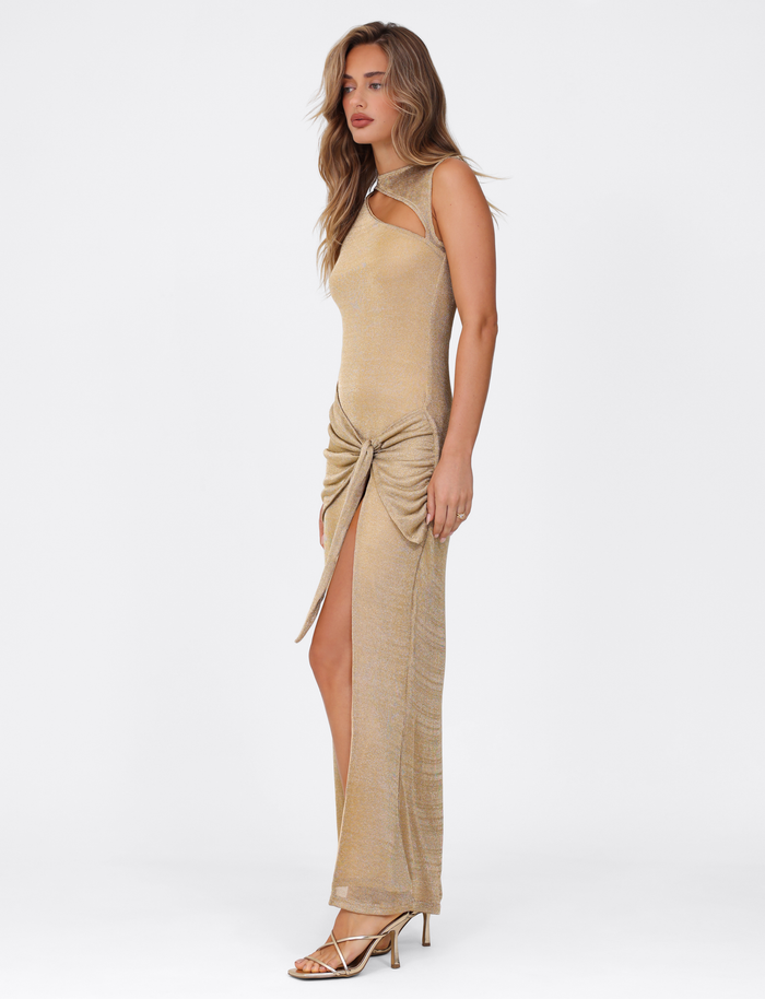 A woman poses against a white background in the TIGERMIST AZELMA MAXI DRESS - GOLD, a sleeveless metallic knit dress with a neckline cutout, ruched detailing, high front slit, and strappy gold heels.