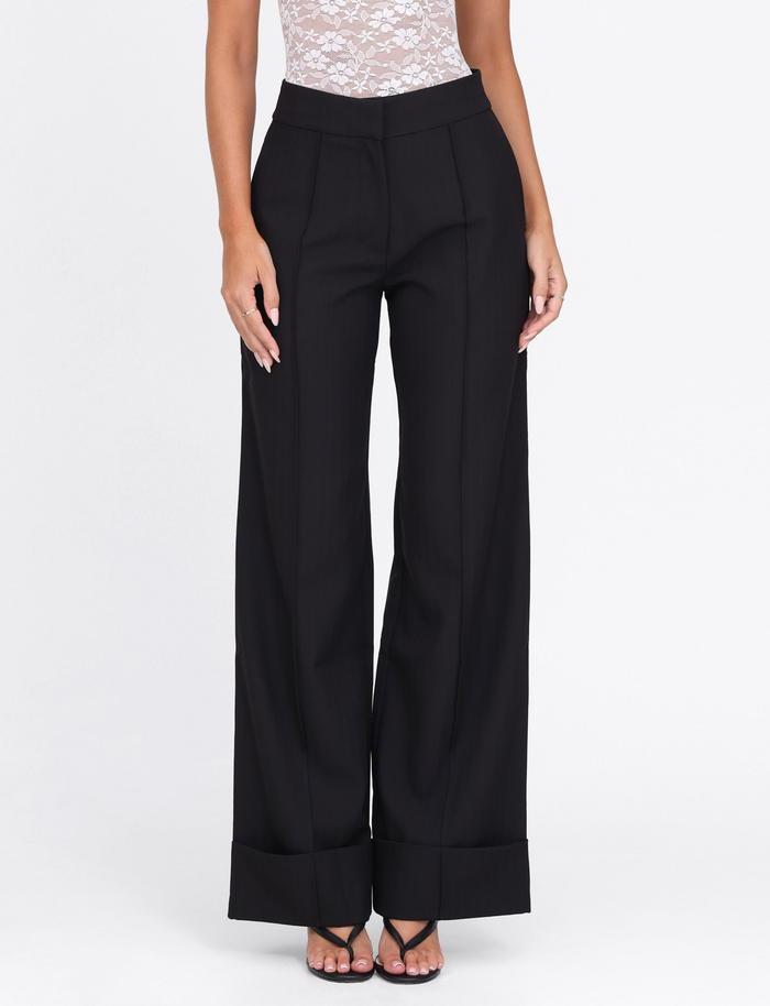 A woman models the Tiger Mist BETHANY PANT in black—high waisted, wide leg with cuffed hems—styled with a sleeveless lace top and strappy black heels, posing against a plain white background.