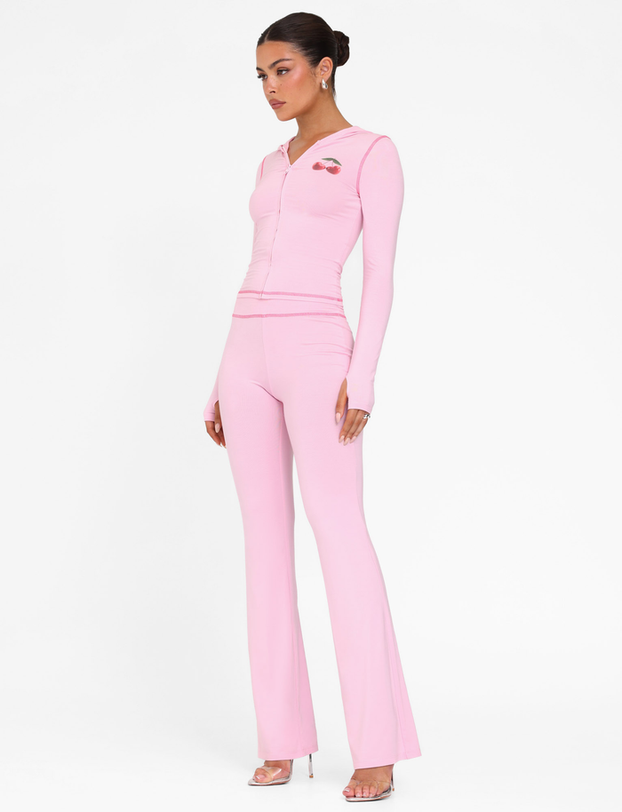 A woman stands against a white background wearing the TIGERMIST BLAYZE PANT in soft pink with a matching fitted zip-up top, her hair styled in a neat bun, and clear high-heeled sandals.