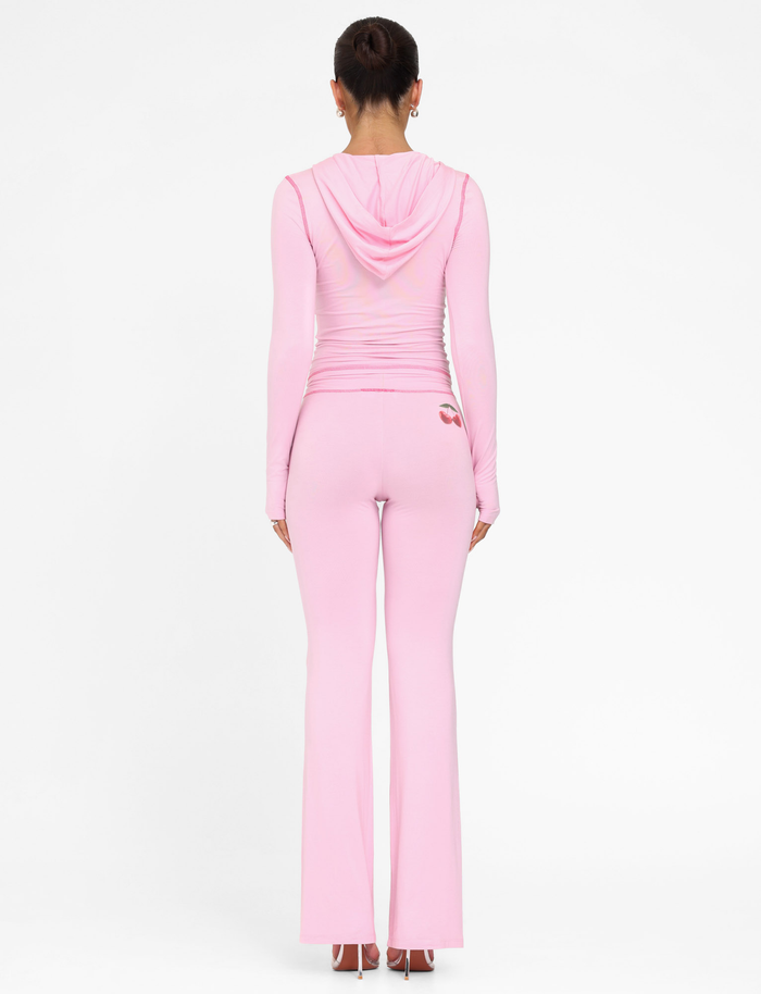 A woman stands facing away, wearing TIGERMIST's BLAYZE PANT in soft pink with a matching fitted light pink hooded top, her hair in a neat bun and arms relaxed by her sides, set against a plain white background.