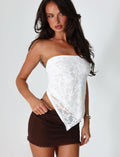 A woman with long dark hair stands against a plain background, wearing a strapless white lace top and a brown mini skirt with ruffled detailing. She has her hands by her sides, adorned with the BRIONY SCARF - WHITE by TIGERMIST, and is looking directly at the camera.