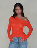 A woman with long dark hair is wearing the BRYNA JUMPER in red by TIGERMIST, which features an off-the-shoulder design and long sleeves with a knit fabrication. She pairs this stylish top with light blue jeans. One arm hangs relaxed by her side while the other rests lightly on her hip against a neutral beige background.
