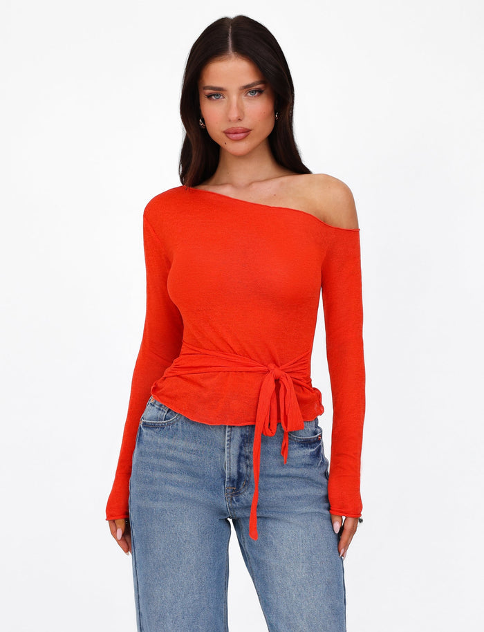 A woman is wearing the BRYNA JUMPER in red by TIGERMIST, featuring an off-the-shoulder design with long sleeves and adjustable styling at the waist, paired with blue jeans. She has long dark hair and is standing against a plain white background.