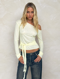 A woman with long blonde hair wears the TIGERMIST BRYNA JUMPER in pale lemon yellow—an off-shoulder, long-sleeve knit sweater with wrap-around ties—paired with dark jeans, standing before a textured beige wall.