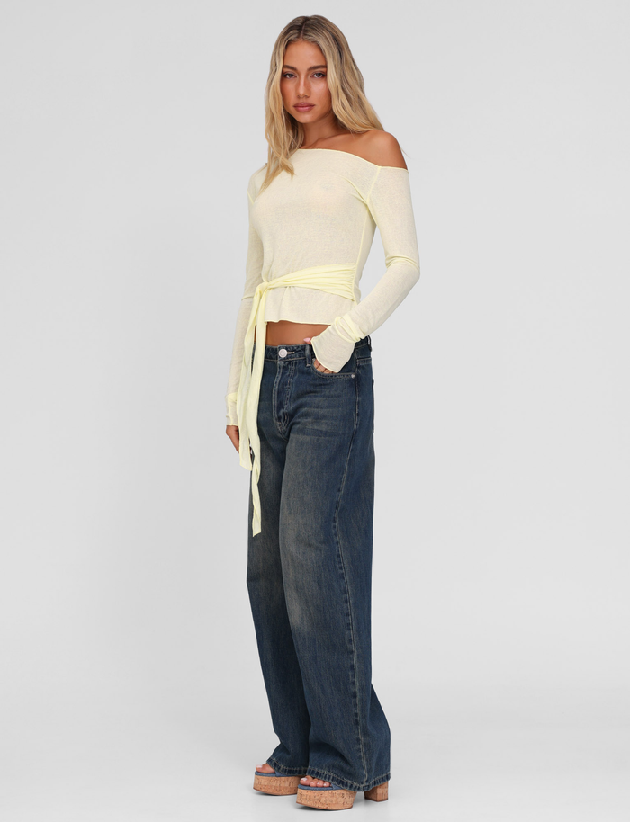 A woman with long blonde hair and a neutral expression stands against a plain background, wearing the TIGERMIST BRYNA JUMPER in pale yellow with wide-leg dark blue jeans and tan platform sandals.
