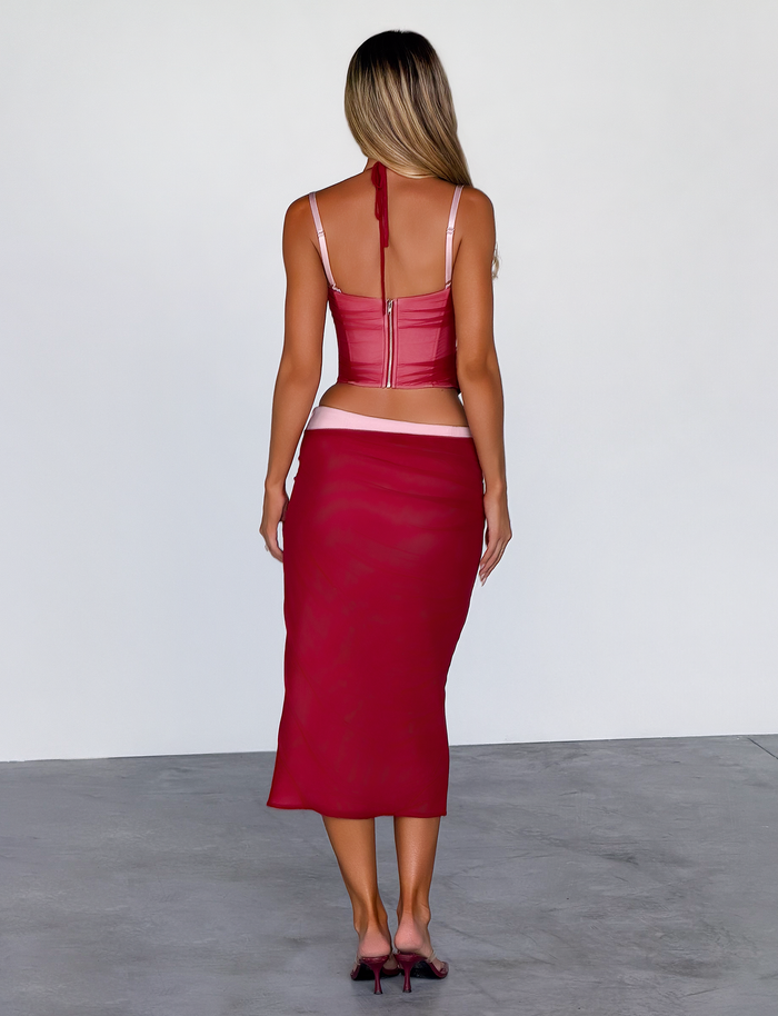 A woman with long, wavy blonde hair faces away, wearing the TIGERMIST CALINE MIDI SKIRT in red with a fitted crop top and matching heels, standing on gray concrete against a plain white wall.