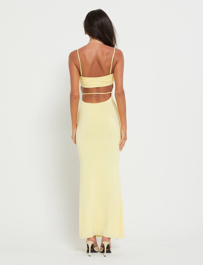 A woman with long dark hair faces away, wearing the TIGERMIST CARA DRESS in pastel citrus yellow—a bodycon bandeau style with cutouts—paired with black high-heeled sandals against a white background.