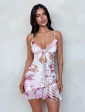 A woman with long dark hair stands against a light-colored wall, showcasing the TIGERMIST CARLY DRESS - MULTI: FLORAL. This sleeveless, ruched dress features a cut-out and tie detail at the front, complemented by a flounced hem. With a neutral expression, she holds her arms relaxed by her sides.