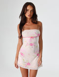 A woman stands against a plain background, wearing the CARSYN MINI DRESS in pink with a watercolor floral design and an asymmetrical hemline by TIGERMIST. Her long dark hair flows over her shoulders as she gazes directly at the camera.