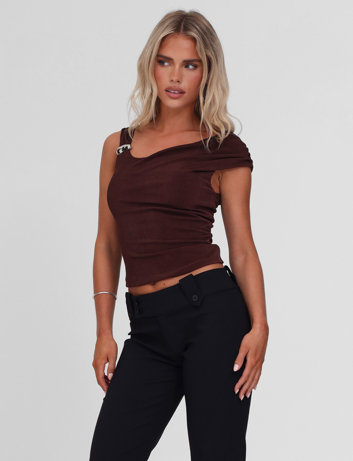 A woman with wavy blonde hair wears the TIGERMIST CASSIAN TOP in chocolate with an off-shoulder detail and silver clasp, paired with high-waisted black pants, standing against a plain light background, looking slightly to the side.